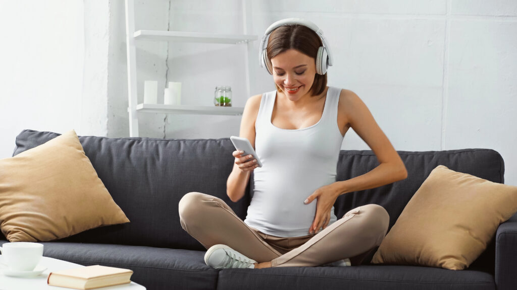 happy pregnant woman sitting on sofa, listening music and chatting on smartphone