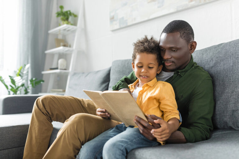 A dad sits on the counch with his young child, as they enjoy a book together.