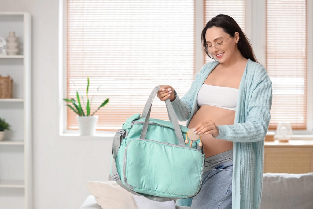 Young pregnant woman putting rattle into maternal hospital bag at home