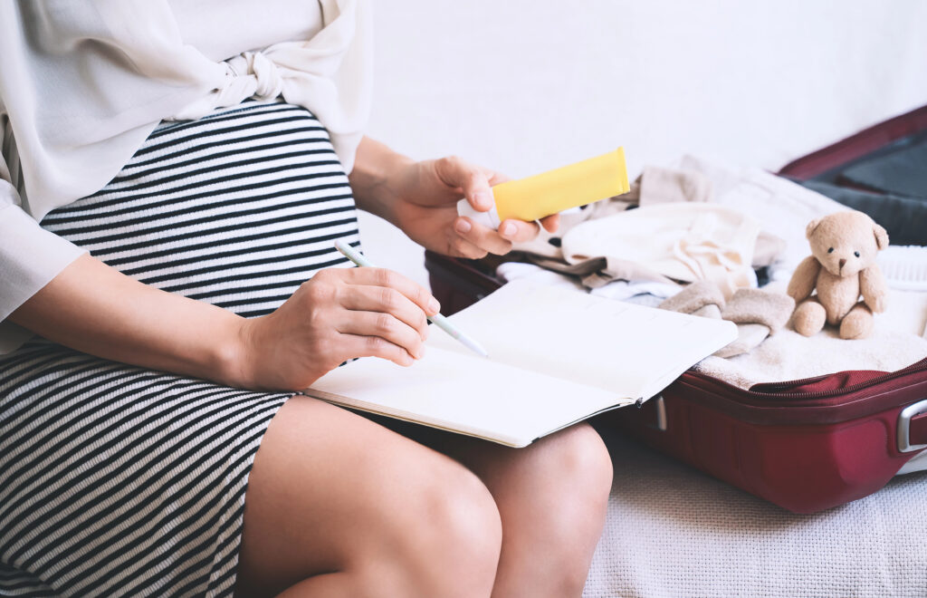 Pregnant woman packing hospital bag with checklist. Expectant mother with travel suitcase of baby clothes and necessities preparing for newborn birth, labor, making notes in paper diary.