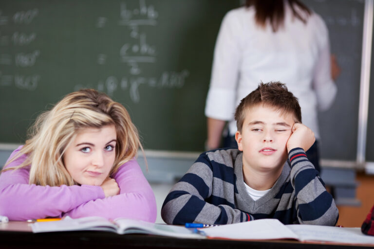 Bored Students Leaning On Desk With Teacher In Background